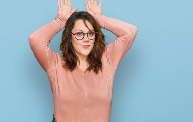 Genç ve cüsseli kadın günlük kıyafetler ve gözlük takıyor. Tavşan kulağı hareketi yapıyor. Avuçlarıyla alaycı ve şüpheci görünüyor. Paskalya tavşanı konsepti. 