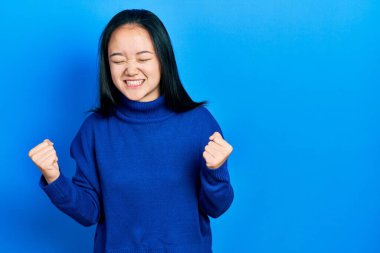 Genç Çinli kız gündelik kıyafetler giyip çok mutlu ve heyecanlı bir şekilde kolları havada, gülümseyen ve başarı için bağıran kazanan bir jest yapıyor. kutlama konsepti. 