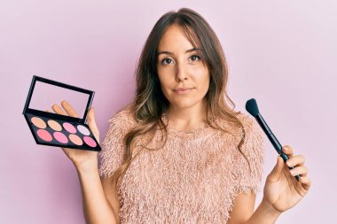 Makyaj fırçası tutan ve yüzünde ciddi bir ifadeyle rahatlamış genç esmer kadın. Basit ve doğal bir şekilde kameraya bakıyorsun.. 