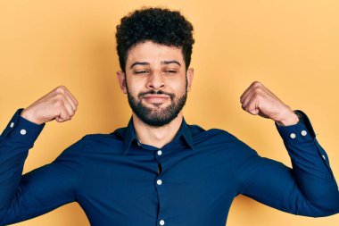 Sakallı, sıradan gömlekli genç bir Arap gururla gülümseyen kol kaslarını gösteriyor. fitness konsepti. 