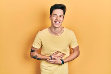 Latin asıllı genç bir adam, günlük sarı tişört giyiyor, gülümsüyor ve kahkahalarla gülüyor.. 
