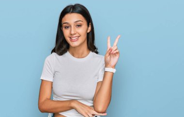 Gündelik beyaz tişört giyen, kameraya gülümseyen ve parmaklarıyla zafer işareti yapan genç İspanyol kız. İki numara.. 