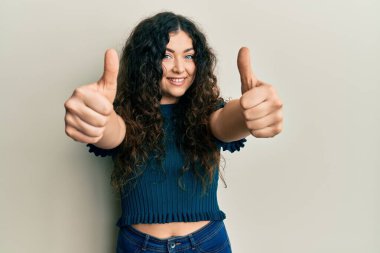 Kıvırcık saçlı, günlük giysiler giyen, el hareketleriyle olumlu jest yapan, gülümseyen ve başarı için mutlu olan genç esmer kadın. kazanan jest. 