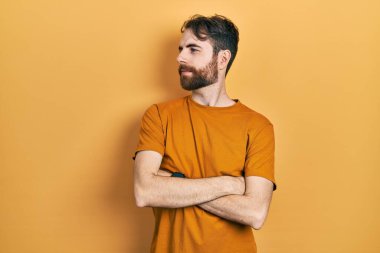 Sakallı, sıradan sarı tişörtlü, kolları çapraz ve kendinden emin beyaz bir adam. 