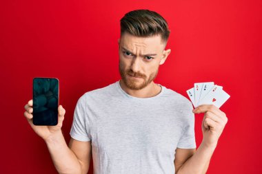 Akıllı telefon ve kumarhane kartı tutan genç kızıl saçlı adam şüpheci ve gergin, sorun yüzünden surat asıyor. negatif kişi. 