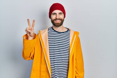 Sakallı, sarı yağmurluklu beyaz adam kameraya bakıp zafer işareti yapıyor. İki numara.. 