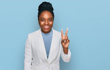 Genç Afro-Amerikan kadın iş kıyafetleri giyip iki numaralı parmaklarıyla işaret ederken kendinden emin ve mutlu bir şekilde gülümsüyor.. 
