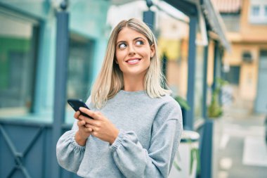 Genç sarışın kız şehirde akıllı telefon kullanarak mutlu bir şekilde gülümsüyor.