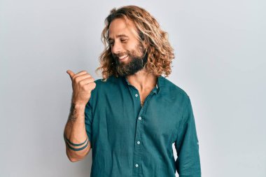 Sakallı, uzun saçlı, sıradan kıyafetler giyen yakışıklı bir adam baş parmağını kaldırıyor ve ağzı açık gülümsüyor. 