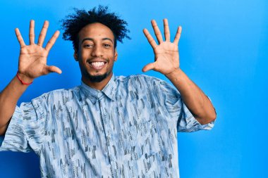 Sakallı, genç, Afro-Amerikan bir adam. Günlük kıyafetlerini giyiyor ve parmaklarıyla on numarayı gösteriyor. Kendinden emin ve mutlu bir şekilde gülümsüyor.. 