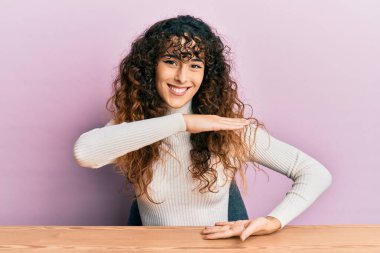 Latin kökenli genç bir kız, gündelik giysiler içinde masaya oturmuş, büyük ve büyük boy işaretleri gösteren el hareketleriyle, ölçü sembolü gösteriyordu. Kameraya bakıp gülümsüyor. ölçüm kavramı. 