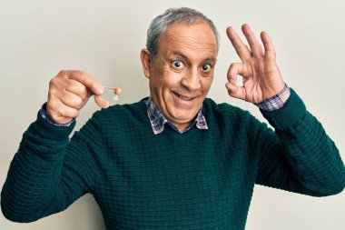 Gri saçlı, elinde tıbbi işitme cihazı olan yakışıklı yaşlı adam parmaklarıyla imza atıyor, gülümsüyor ve mükemmel bir sembol oluşturuyor. 