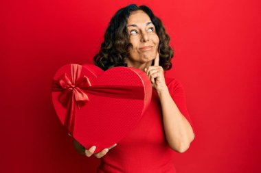 Orta yaşlı İspanyol bir kadın sevgililer günü hediyesi tutuyor ciddi bir yüz ifadesi var eli çenesinde soruyla ilgili düşünüyor, kafa karıştırıcı bir fikir hakkında düşünceli. 