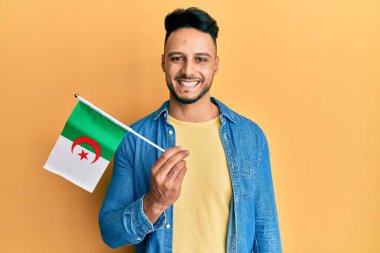 Cezayir bayrağını tutan genç Arap adam pozitif ve mutlu görünüyor. Kendine güvenen bir gülümsemeyle ayakta duruyor ve dişlerini gösteriyor. 