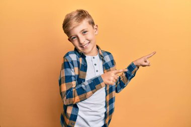 Kafkasyalı küçük çocuk günlük kıyafetler giymiş gülümseyen ve kameraya bakıp iki eliyle ve parmaklarıyla kenarı işaret ediyor.. 