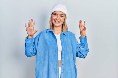 Güzel sarışın kadın yün şapka takıyor ve 8 numaralı parmaklarıyla işaret ederken kendinden emin ve mutlu bir şekilde gülümsüyor.. 