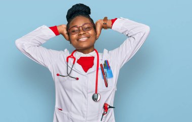 Genç Afrikalı Amerikalı kadın doktor üniforması giyiyor ve steteskop takıyor. Rahatlıyor ve esniyor, kolları ve elleri başının arkasında ve boynunun arkasında mutlu bir şekilde gülümsüyor. 