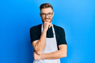 Profesyonel önlük giymiş genç kızıl saçlı adam kameraya kollarını kavuşturmuş ve çenesi üzerinde kendinden emin bir şekilde bakıyor. Olumlu düşünmek. 