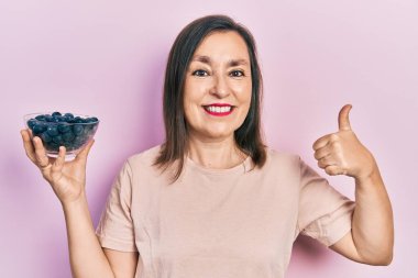 Orta yaşlı, elinde böğürtlen tutan, mutlu ve pozitif gülümseyen, başparmağıyla mükemmel ve onay işareti yapan İspanyol bir kadın. 