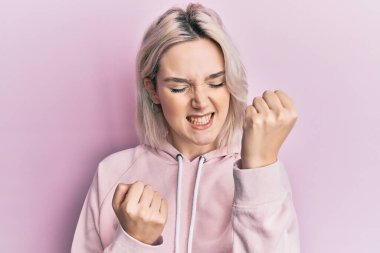 Gündelik giysiler giyen genç sarışın kız şaşkınlık ve şaşkınlık içinde kollarını kaldırarak ve gözlerini kapayarak başarıyı kutluyor. 