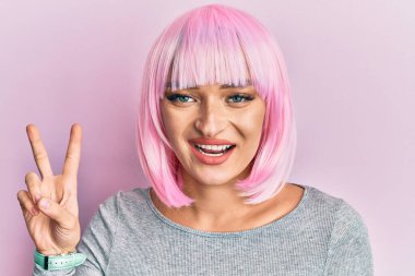 Pembe peruklu genç beyaz kadın kameraya bakıp gülümsüyor ve zafer işareti yapıyor. İki numara.. 