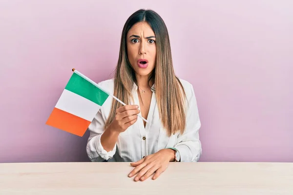 Jovem Hispânica Segurando Bandeira Irlandesa Sentada Mesa Assustada Espantada Com — Fotografia de Stock