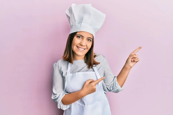 年轻美丽的女人穿着职业厨师的制服 戴着帽子 面带微笑 看着摄像机 用双手和手指指向旁边 — 图库照片