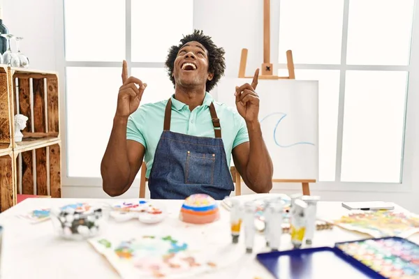 年轻的非洲裔美国人坐在艺术工作室的桌子上 惊讶地抬起头 用手指和胳膊指指着 — 图库照片