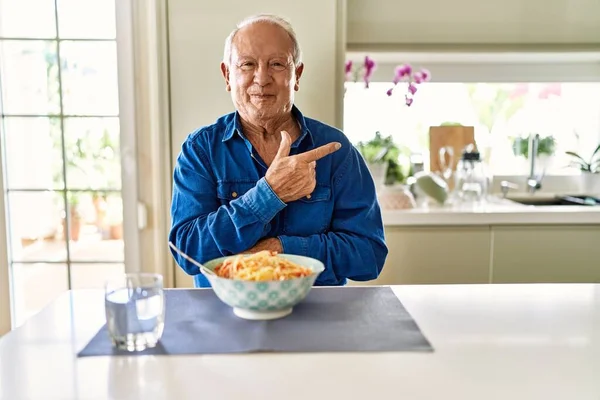 Senior man with grey hair eating pasta spaghetti at home cheerful with a smile of face pointing with hand and finger up to the side with happy and natural expression on face