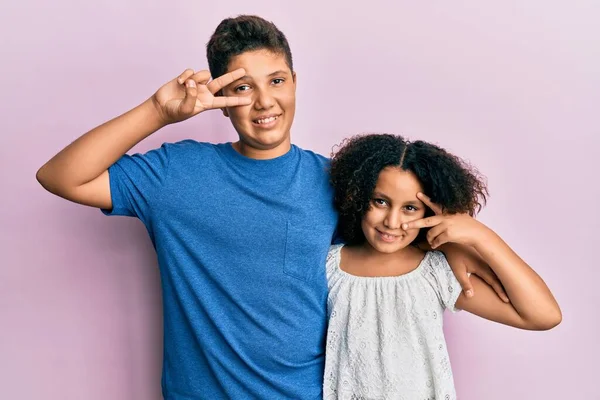 顔の上に指で平和の象徴を一緒にカジュアルな服を着て弟と妹の若いヒスパニック系の家族 勝利を示す明るい笑顔 — ストック写真