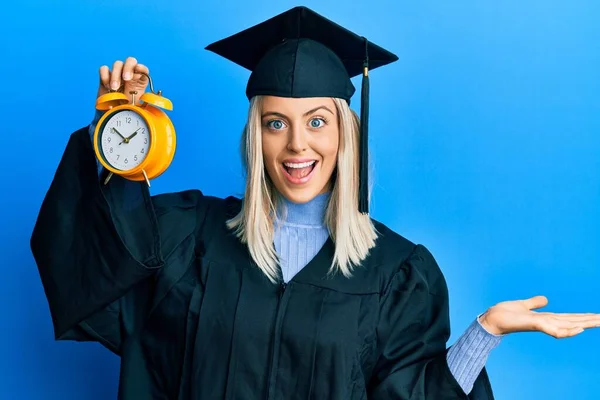 卒業キャップと式のローブを身に着けている美しいブロンドの女性は幸せな笑顔で勝利を祝う目覚まし時計を保持します — ストック写真