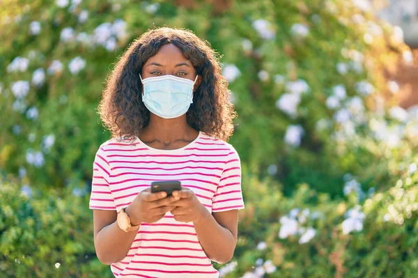 Νεαρή Αφροαμερικανή Που Φοράει Ιατρική Μάσκα Χρησιμοποιώντας Smartphone Στο Πάρκο — Φωτογραφία Αρχείου