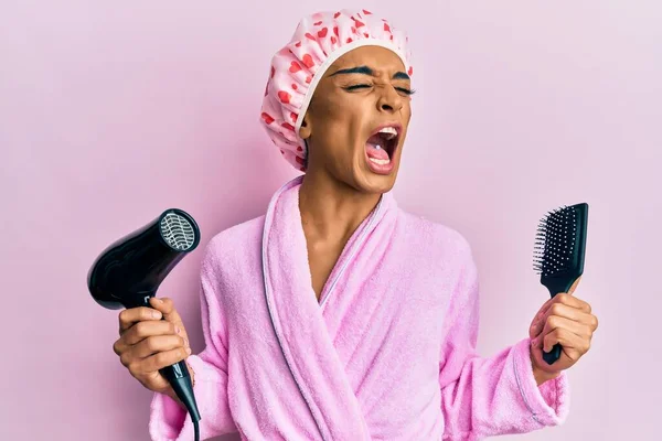 Hispanic man wearing make up wearing shower cap holding hair dryer and comb angry and mad screaming frustrated and furious, shouting with anger. rage and aggressive concept.