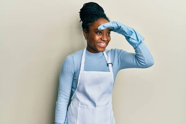 African american woman with braided hair wearing cleaner apron and gloves very happy and smiling looking far away with hand over head. searching concept.