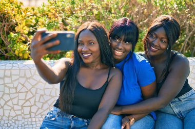 Üç Afrikalı Amerikalı arkadaş mutlu bir şekilde gülümsüyor. Parktaki akıllı telefonun yanında selfie çekiyorlar..