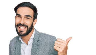 İş kıyafetleri giymiş genç İspanyol bir adam arka tarafı işaret ediyor. Elleri havada, kendinden emin bir şekilde gülümsüyor. 