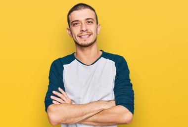 Gündelik kıyafetler giyen İspanyol genç adam kameraya bakarken kollarını kavuşturup gülümsüyor. pozitif kişi. 