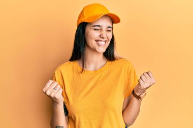 Genç İspanyol kadın teslimat üniforması giyiyor ve çok mutlu ve heyecanlı kollarını kaldırıp, gülümseyerek ve başarı için bağırarak kazanan jestler yapıyor. kutlama konsepti. 