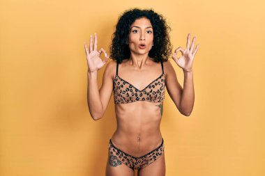 Bikini giyen genç Latin kız şaşırmış ve şaşırmış görünüyor. Tamam sembolünü parmaklarıyla yapıyor. çılgın ifade 