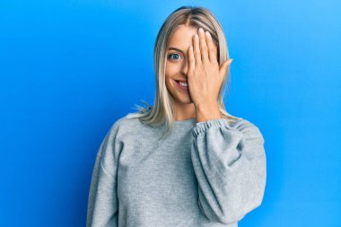 Güzel sarışın kadın gündelik kıyafetler giyiyor. Bir gözünü eliyle kapatıyor. Yüzünde kendine güvenen bir gülümseme ve sürpriz bir duygu var.. 