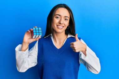 Genç İspanyol kız, neşeli ve pozitif gülümseyen renkli bulmaca küp zeka oyunu oynuyor, başparmağıyla mükemmel ve onay işareti yapıyor. 