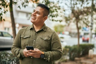 Genç Latin adam şehirde akıllı telefon kullanarak mutlu bir şekilde gülümsüyor..