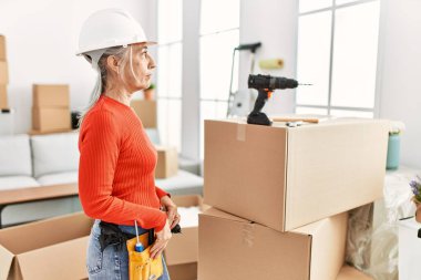 Orta yaşlı, gri saçlı bir kadın yeni evinde dikilip yan yana bakıyor, rahat profil pozu veriyor, kendine güvenen bir gülümsemesi var.. 