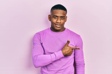 Genç siyah adam, günlük pembe süveter giyiyor. Neşeli, yüzünde bir gülümseme var. Mutlu ve doğal bir ifadeyle elini ve parmağını kaldırıyor. 