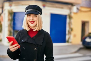 Genç sarışın kadın şehirde akıllı telefon kullanarak mutlu bir şekilde gülümsüyor..