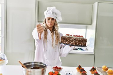 Beyaz tenli pasta şefi kadın mutfakta pasta ve pasta pişiriyor. Kızgın suratlı, olumsuz işaretli, baş parmakları aşağıda, reddedilme kavramından hoşlanmadığını gösteriyor. 