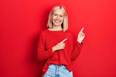 Güzel sarışın kadın, sıradan kırmızı bir süveter giyiyor gülümsüyor ve kameraya bakıyor iki eliyle ve parmaklarıyla kenarı işaret ediyor.. 