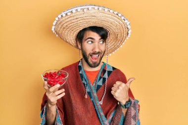 Meksikalı şapkalı genç İspanyol adam elinde kırmızı biber, baş parmağını kaldırıyor ve ağzı açık gülümsüyor. 