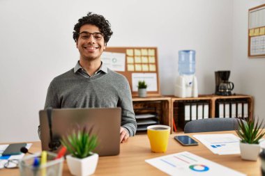 Ofisteki masasında oturan, yüzünde mutlu ve havalı bir gülümseme olan, iş tarzı giyinmiş genç İspanyol bir adam. şanslı kişi. 
