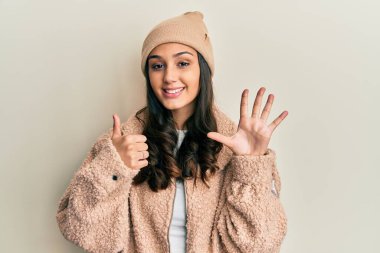 Genç İspanyol kadın yün kazak ve kış şapkası giyer ve altı numaralı parmaklarıyla işaret ederken kendinden emin ve mutlu bir şekilde gülümser.. 
